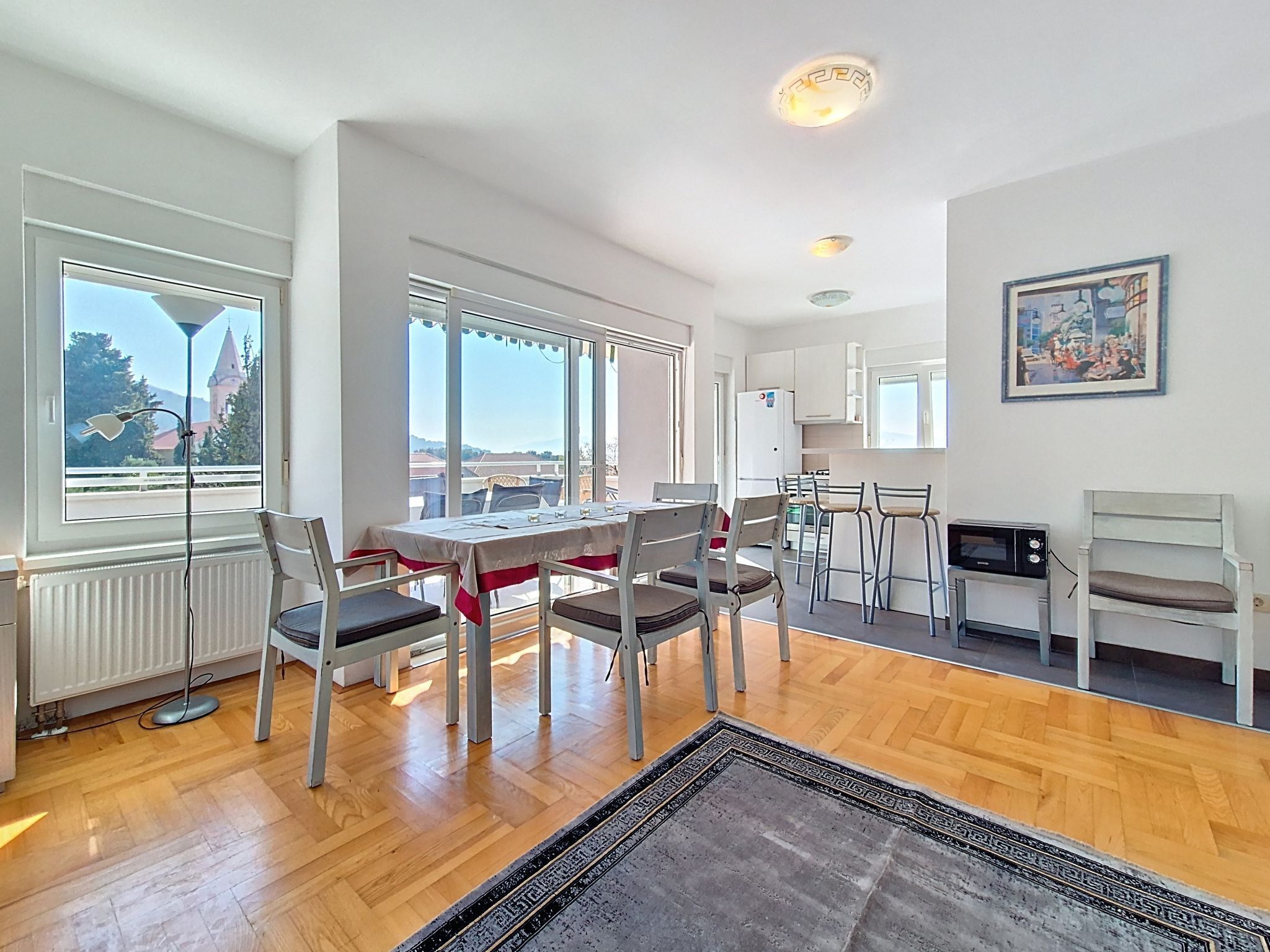 Uma sala de jantar com mesa e cadeiras, uma cozinha com micro-ondas e vista para um edifício através de grandes janelas.