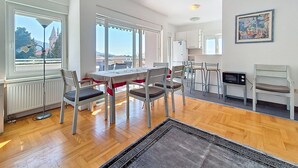 A dining area with a table and chairs, a kitchen with a microwave, and a view of a building through large windows. - Apartment Rilić by Interhome (Zaostrog)