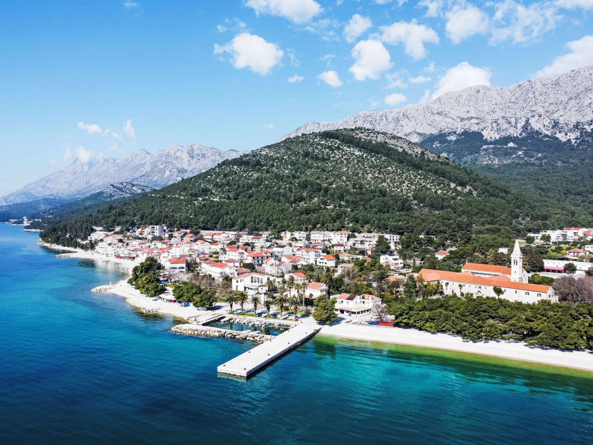 A coastal town with a pier, clear blue waters, and a mountainous backdrop.
