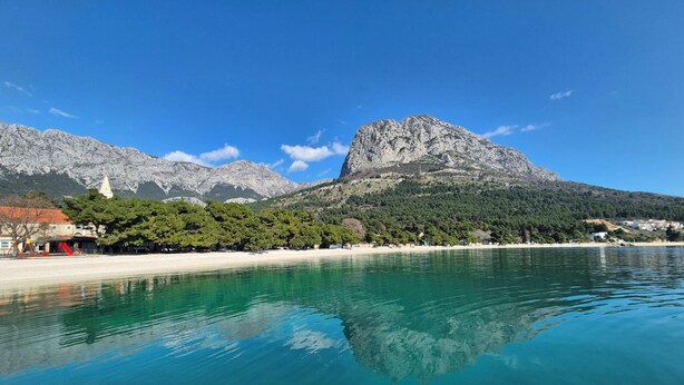 A coastal town with clear turquoise waters, a sandy beach, and a prominent mountain in the background.