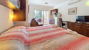 A modern hotel room with a bed, a TV, a kitchenette, and a sliding glass door to the outside.