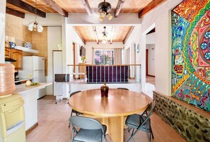A dining area with a round table, chairs, a ceiling fan, a kitchen with a refrigerator, and a colorful mural on the wall.