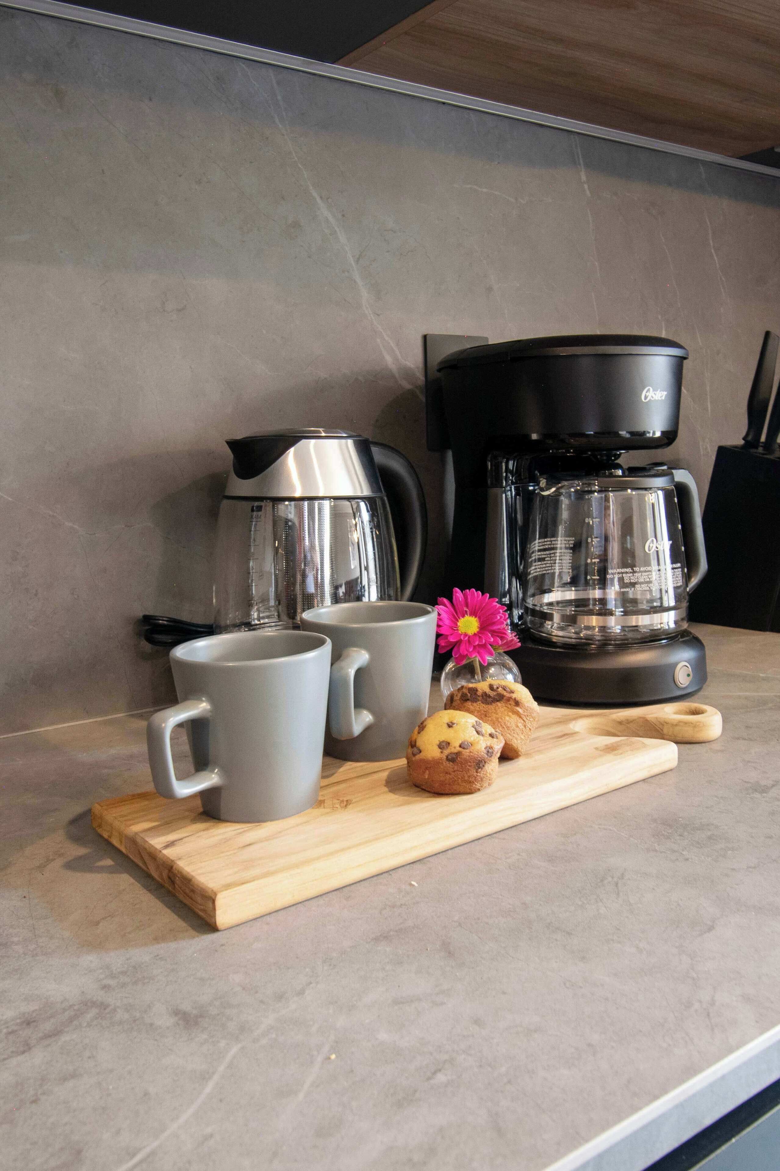 Un comptoir de cuisine avec une machine à café, deux tasses, un muffin et une fleur sur une planche en bois.