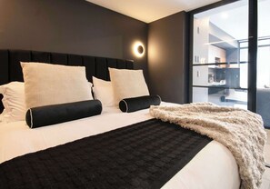 Ein modernes Hotelzimmer mit einem großen Bett, einer runden Wandleuchte und Blick auf die Küche durch eine Glastür.