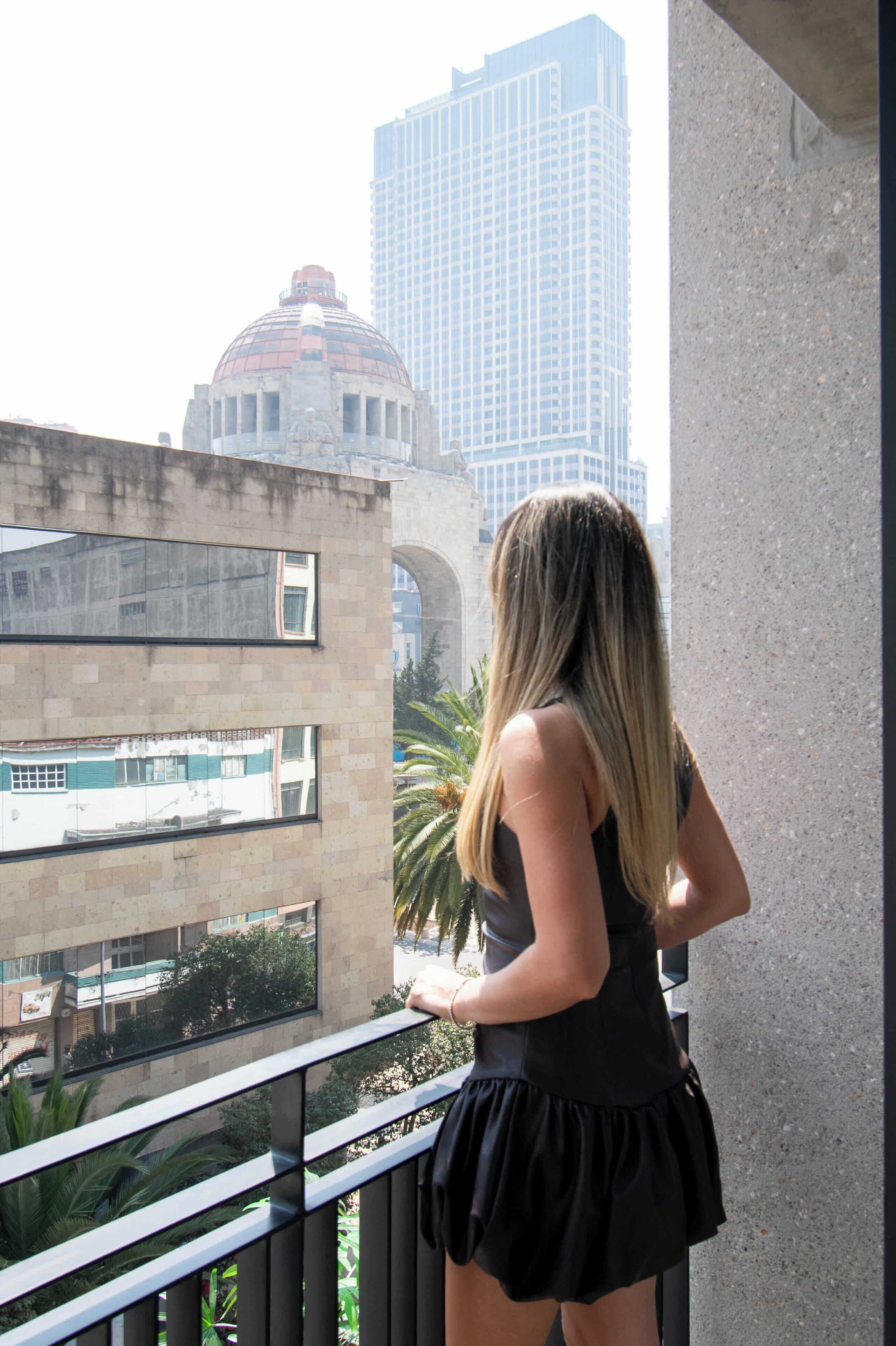 Uma pessoa em uma varanda com vista para uma paisagem urbana com edifícios e uma estrutura em forma de cúpula.