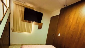 Ein Schlafzimmer mit einem Bett, einem Fenster mit Jalousien, einem an der Wand montierten Fernseher und einer Holztür.