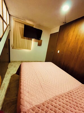 Ein Schlafzimmer mit einem Bett, einem Fenster mit Jalousien, einem an der Wand montierten Fernseher und einer Holztür.