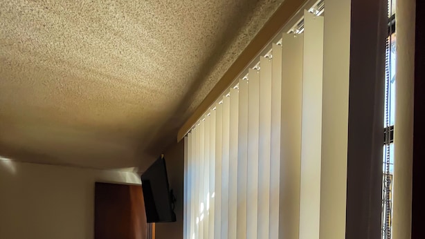 Ein Zimmer mit einem Bett, einem an der Wand befestigten Fernseher und einem Fenster mit vertikalen Jalousien.