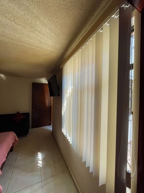 Una stanza con un letto, una televisione a muro e una finestra con persiane verticali.