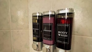 Ein an der Wand befestigter Spender mit drei Flaschen, beschriftet mit 'Shampoo', 'Conditioner' und 'Body Wash'.