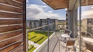 Uma varanda com vista para um complexo de edifícios modernos, guardrails de vidro e uma pequena mesa com uma cadeira.