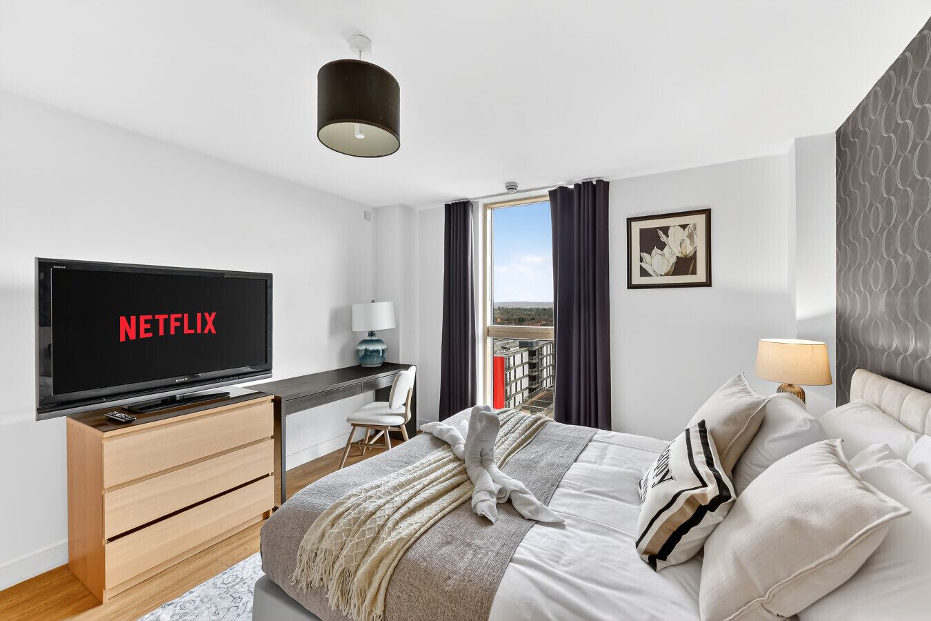 A modern hotel room with a bed, a desk, a television displaying 'NETFLIX,' a window with a view, and a patterned wall.