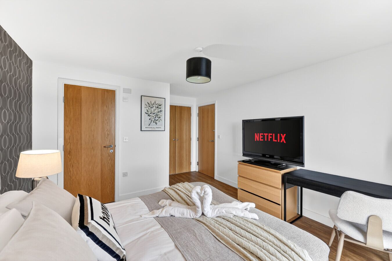 Una camera d'albergo con un letto, una scrivania con una TV che mostra 'NETFLIX', una lampada e un quadro botanico appeso al muro.