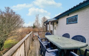A wooden deck with a table and chairs, a white building with a blue window, and leafless trees in the background. - Stunning home in årred with WiFi (Årred)