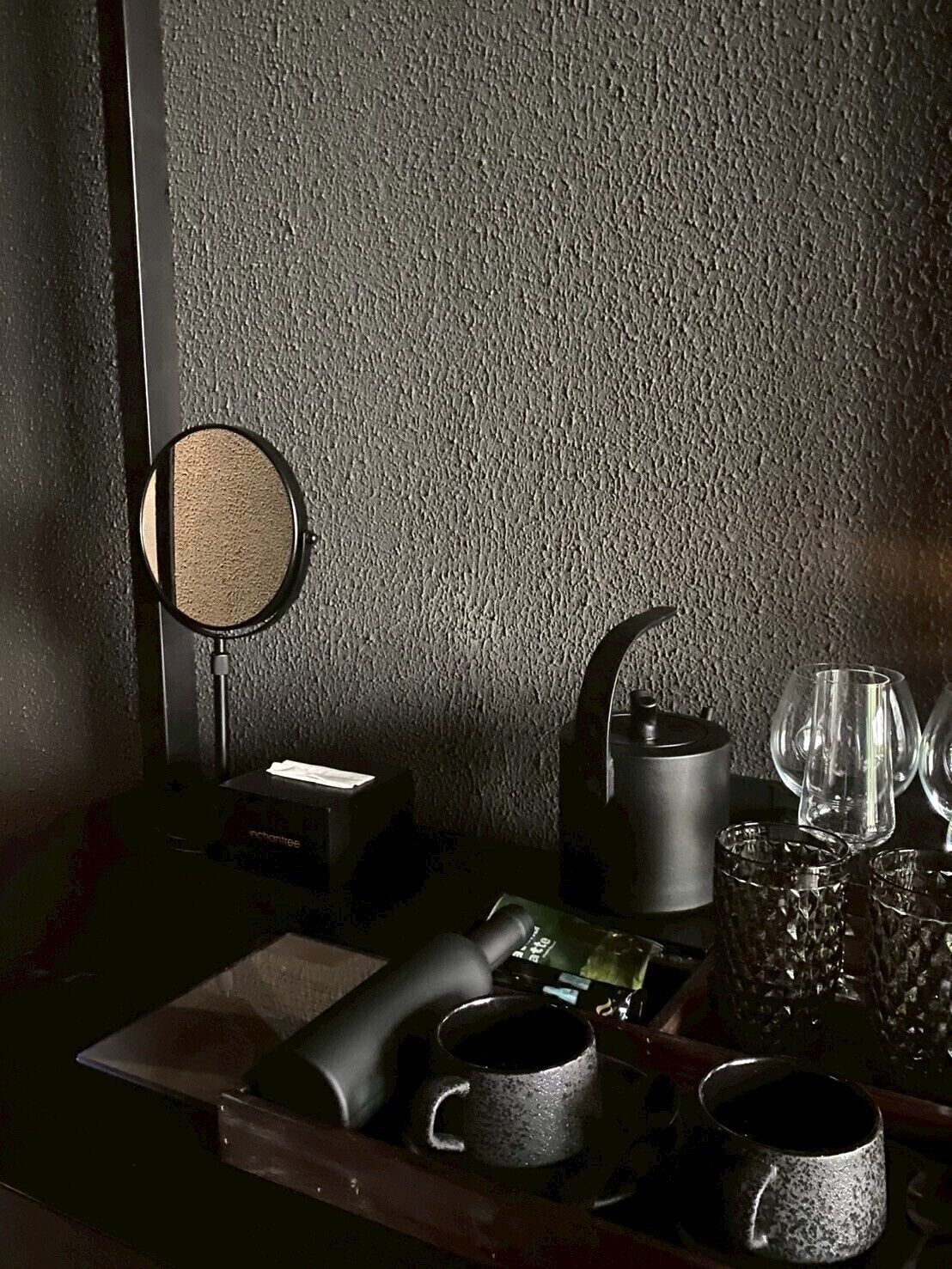 A dimly lit room with a textured wall, a round mirror, a small table with a teapot, cups, and glasses.