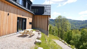 A modern building with wooden siding, a patio with a table and benches, and a view of a forested valley. - Chalet in a beautiful setting (Albrechtice v Jizerskych horach)