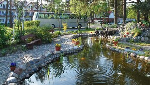 Ein Teich mit einer Springbrunnen, umgeben von Steinen und Kübelpflanzen, mit einem Bus und Gebäuden im Hintergrund.