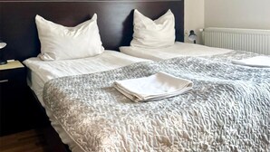 Ein Hotelzimmer mit zwei Betten, weißen Leinentextilien, einem dunklen Kopfteil, einem Nachttisch mit einer Lampe und einer Heizung.