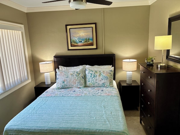 Une chambre à coucher avec un lit, deux lampes de chevet, une commode, un ventilateur de plafond, une fenêtre avec des stores et un tableau encadré au mur.