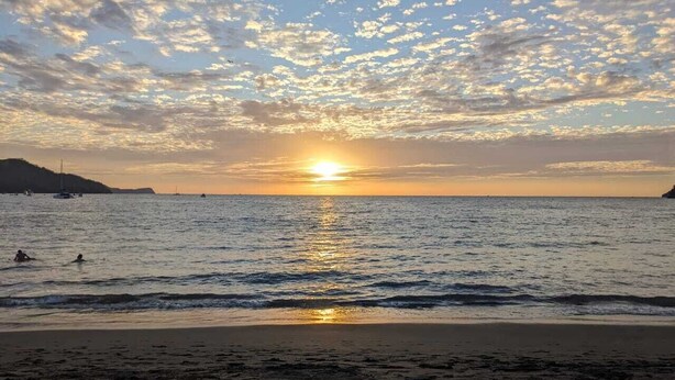 Une plage où le soleil se couche sur l’eau, des gens qui se baignent et des montagnes qui se dessinent à l’horizon.