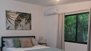 Une chambre à coucher comprenant un lit, une table de chevet, un appareil de climatisation, une fenêtre avec des rideaux et un tableau accroché au mur.