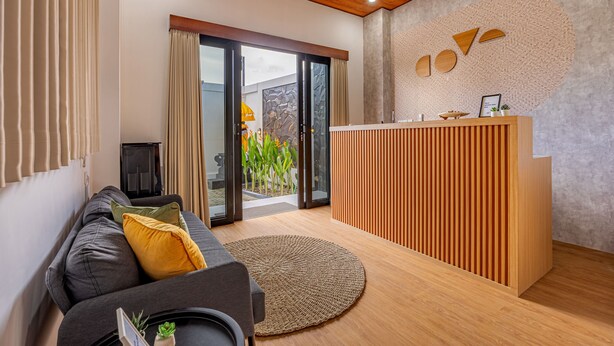 A modern living room with a sofa, a round rug, a wooden reception desk, and a sliding glass door leading to an outdoor area. - Cove Abasan (Tibubeneng)