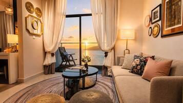A living room with a view of the sunset over the water, a sofa, a chair, a coffee table, and a lamp.