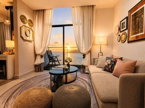 Un salon avec vue sur le coucher de soleil sur l’eau, comprenant un canapé, un fauteuil, une table basse et une lampe.