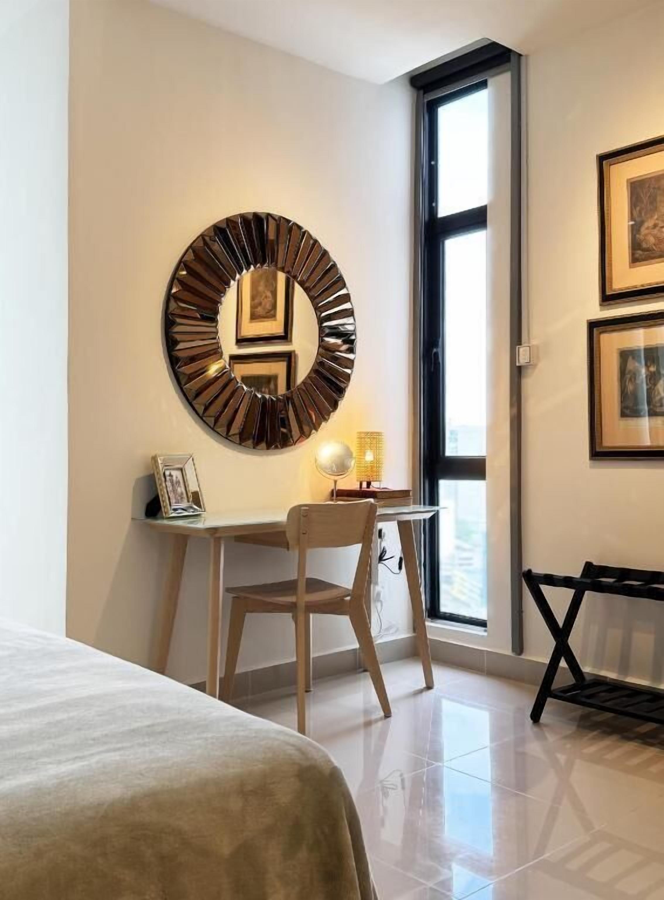 Ein modernes Schlafzimmer mit einem Bett, einem Schreibtisch mit Stuhl, einem runden Wandspiegel und gerahmten Bildern.