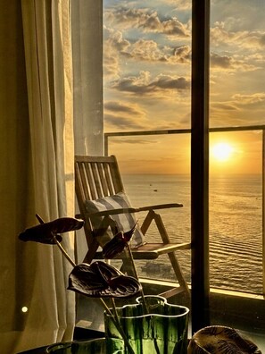 Une pièce avec vue sur la mer au coucher du soleil, une chaise, une table et une plante.