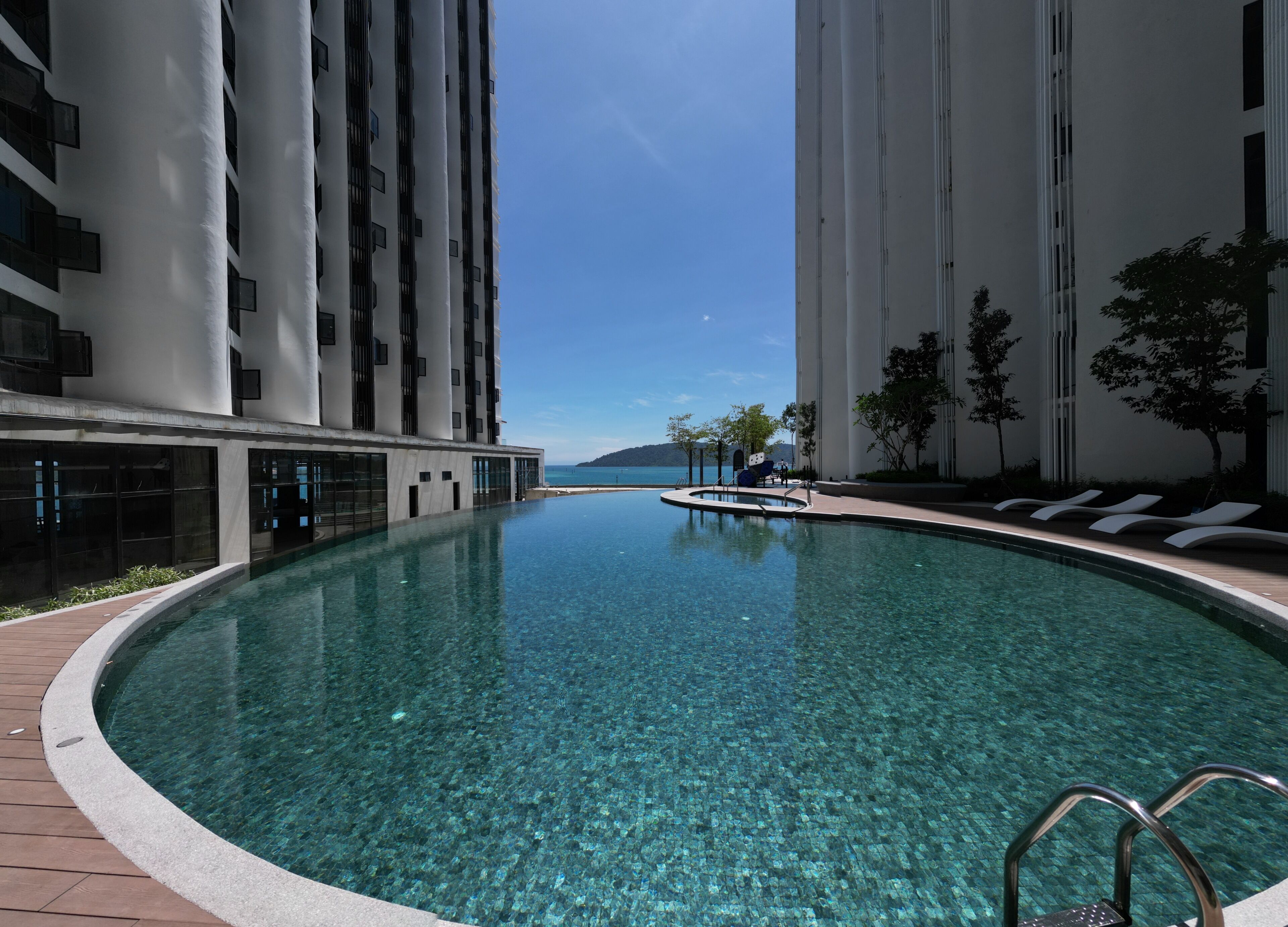 A swimming pool with clear water, surrounded by modern buildings, and a view of the sea.