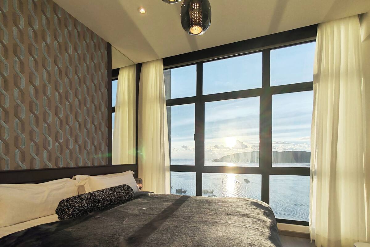 Een slaapkamer met een groot bed, een behang met patroon, een raam met gordijnen en uitzicht op een waterpartij.