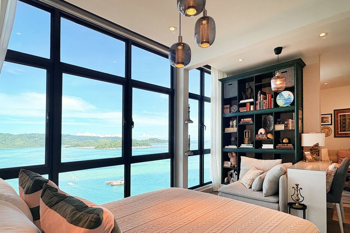 Een slaapkamer met een groot raam, een bed, een boekenkast en uitzicht op zee.