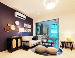 Een moderne woonkamer met een bank, een salontafel en een lamp. Er is een luchtverkoeler aan de muur en er hangen afbeeldingen in lijsten.
