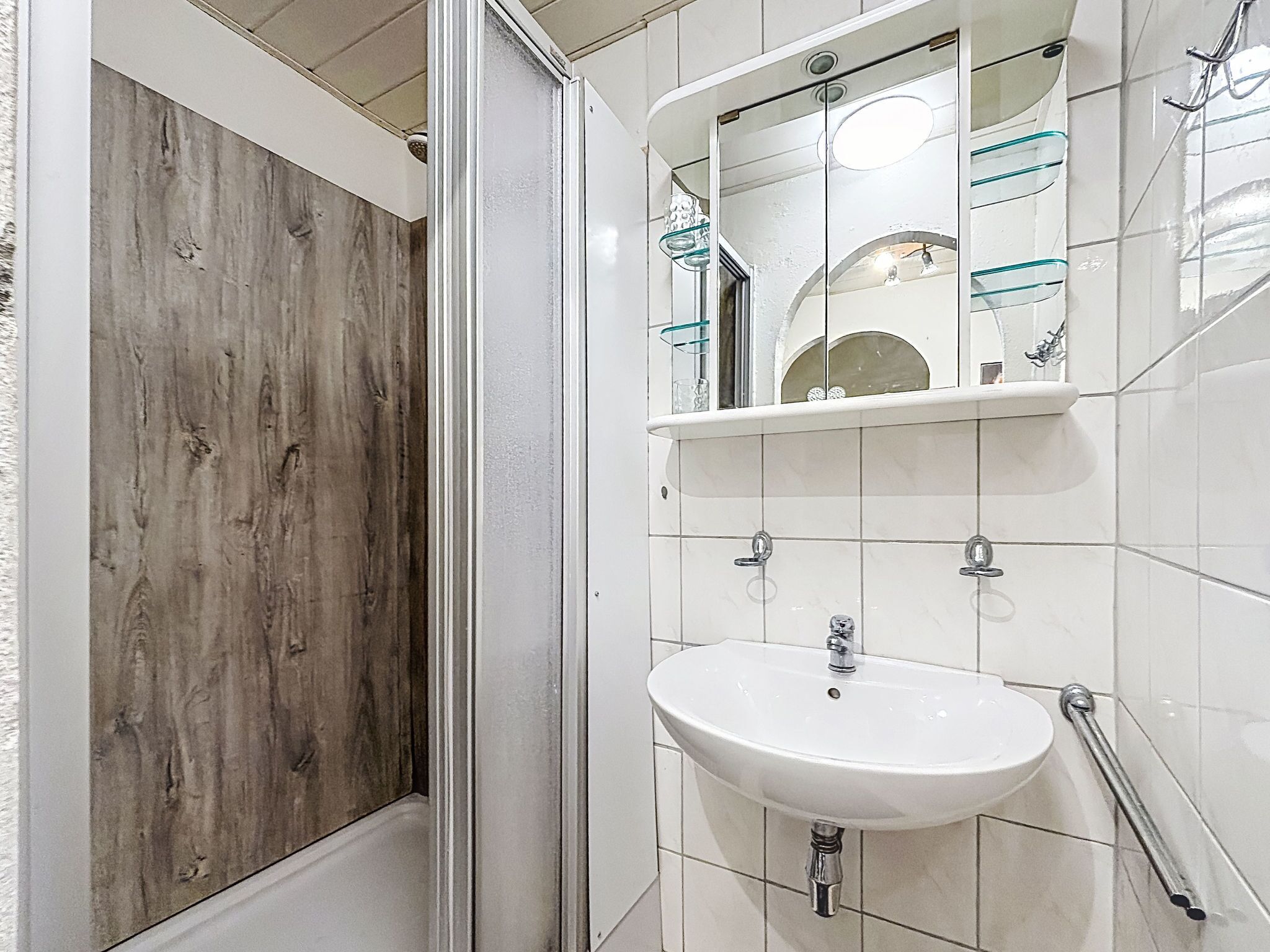 Une salle de bain avec un espace de douche en bois, un lavabo blanc avec un miroir au-dessus, et une étagère en verre où sont rangés les articles de toilette.