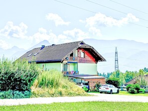 Ein zweistöckiges Haus mit Balkon, umgeben von Grünflächen und parkenden Autos.