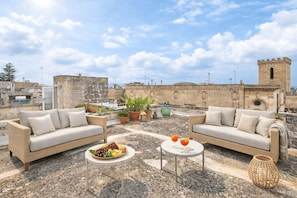 Una terrazza sul tetto con due divani, un tavolo con frutta e bevande, piante in vaso e un edificio storico con una torre sullo sfondo.