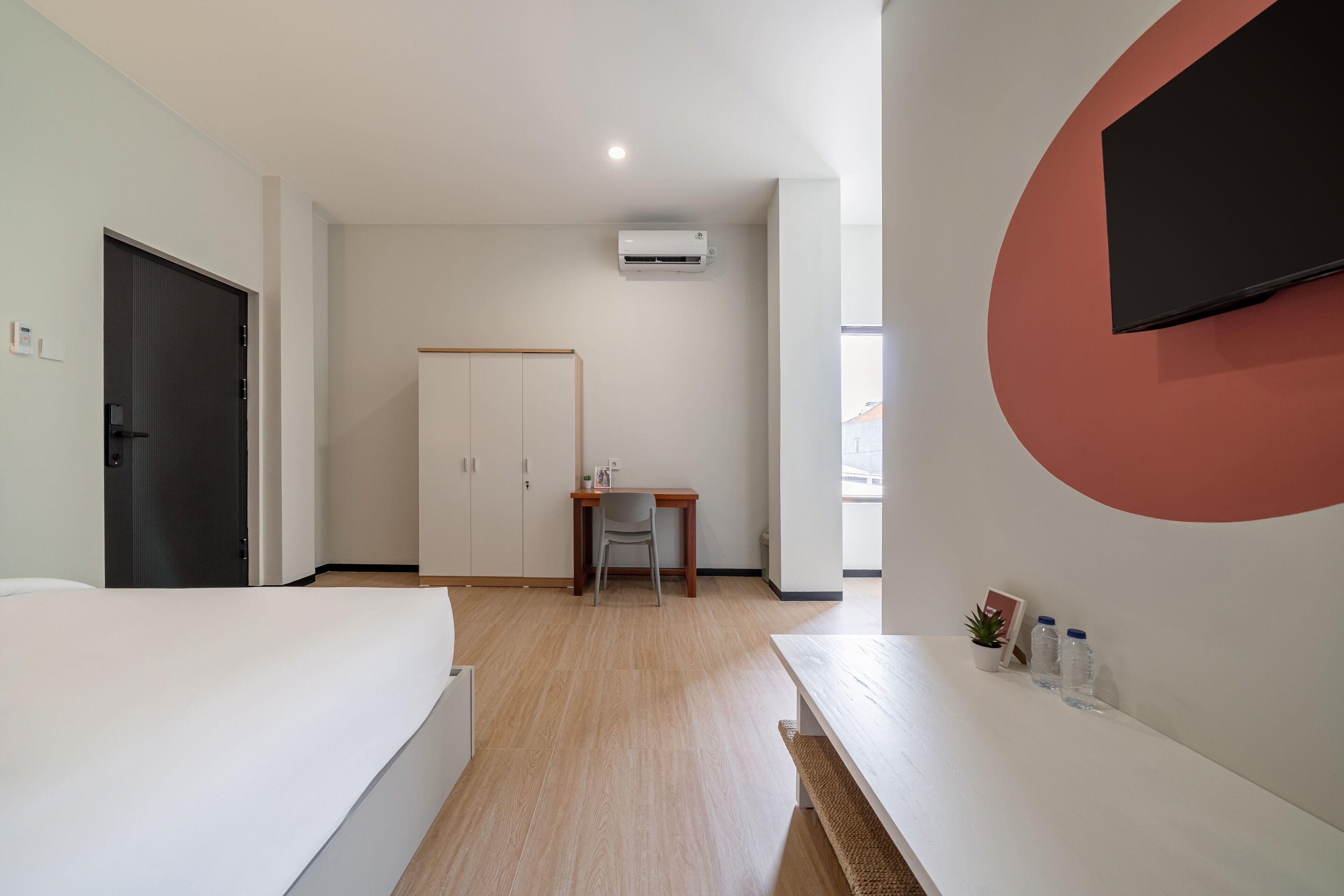 A hotel room with a bed, a desk, a chair, a TV, and an air conditioning unit.