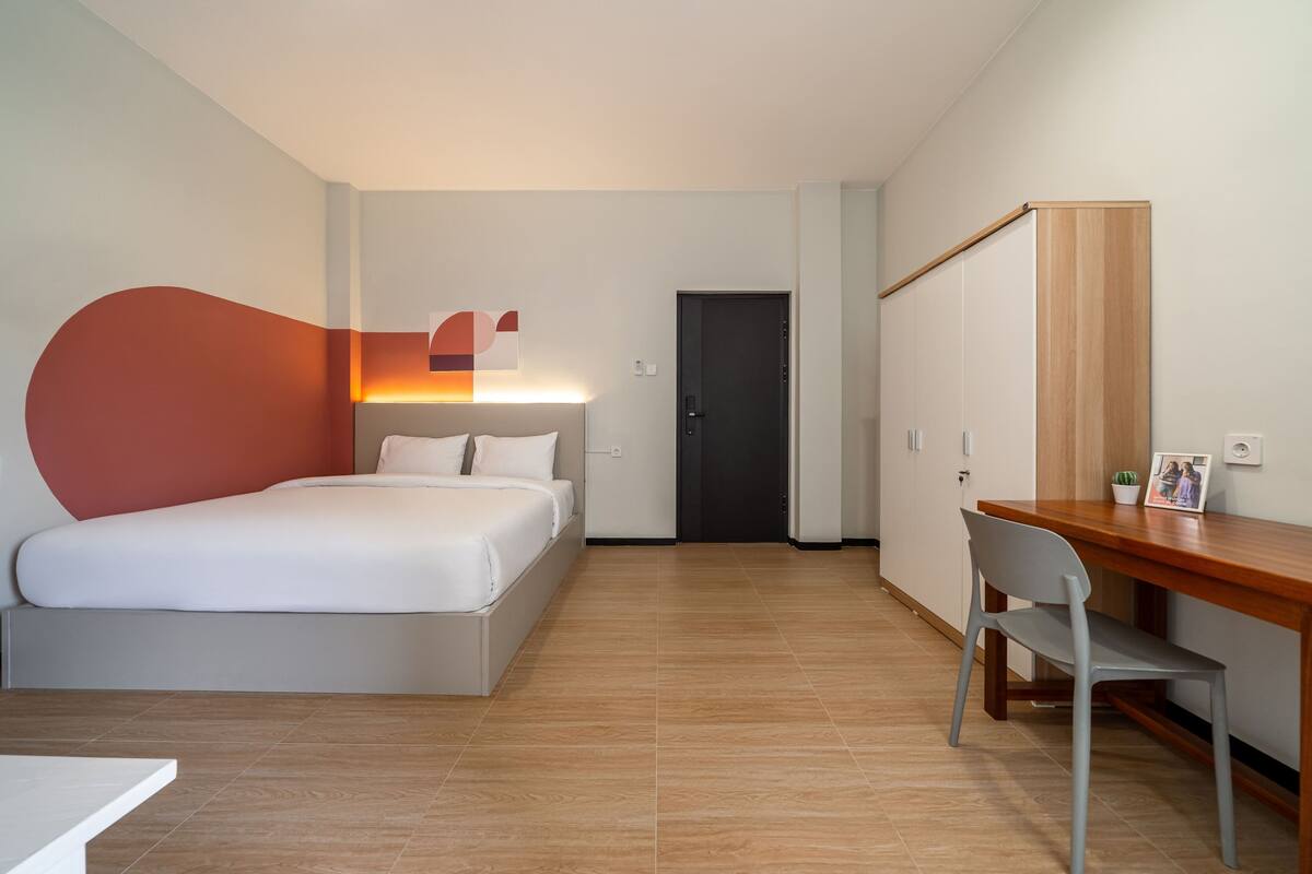 A modern hotel room with a bed, bedside table, desk, chair, wardrobe, and a door.