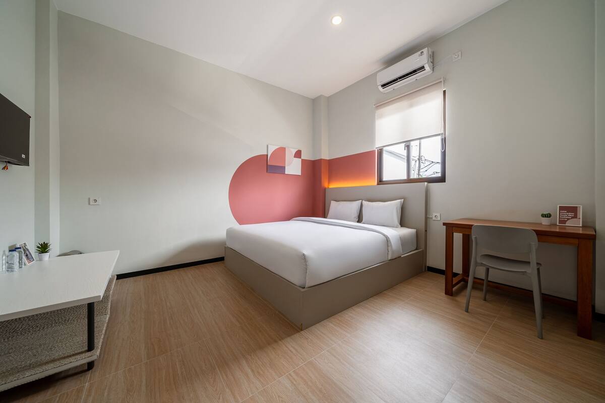A modern hotel room with a bed, desk, and chair, a small table with a plant, and a wall-mounted air conditioner.
