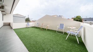 Terraza en la azotea con césped artificial, una pared blanca y algunas sillas y mesas.