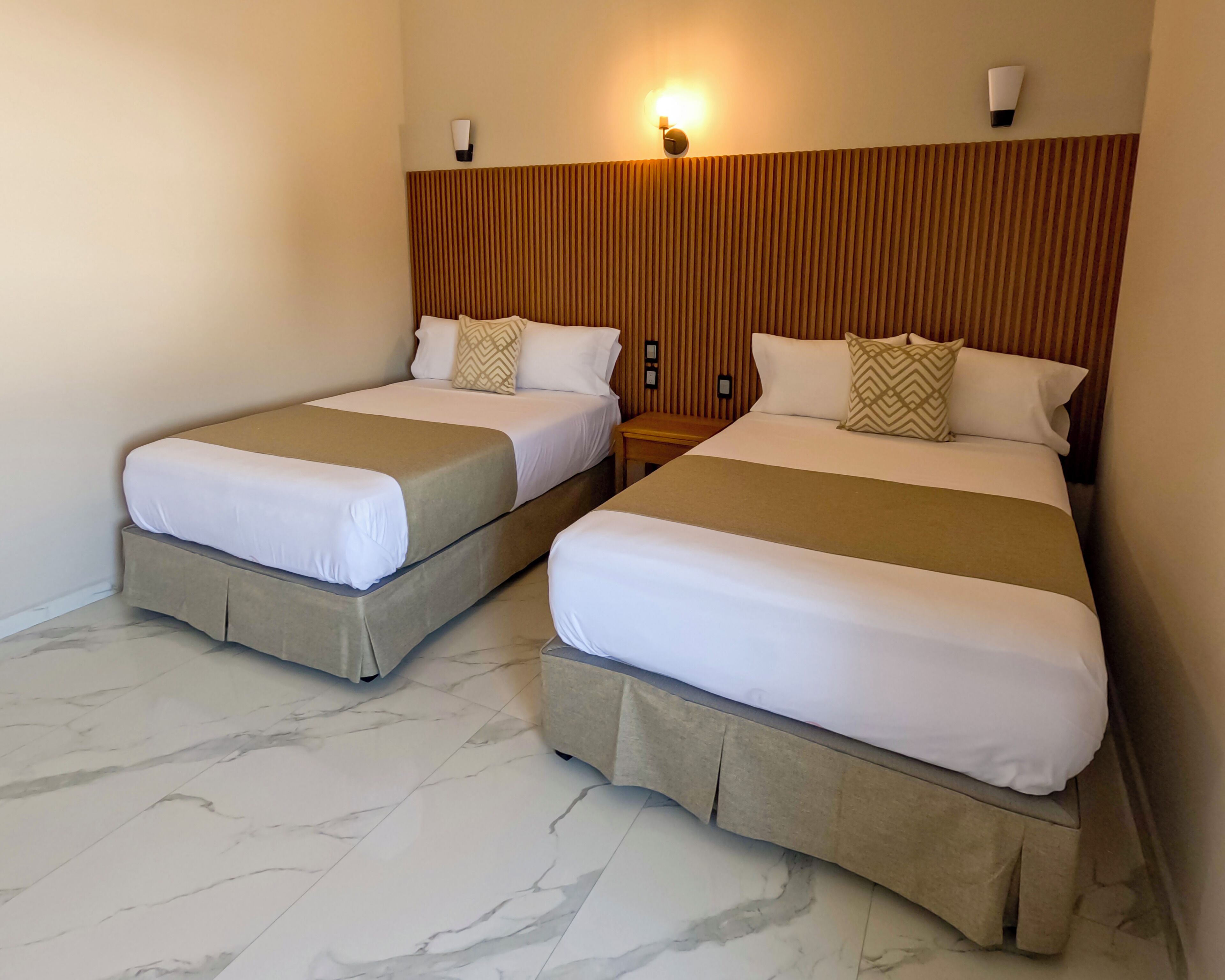 Two twin beds with beige bedding, a wooden headboard, and a wall-mounted light fixture.