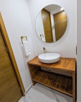 Un baño con espejo redondo, lavamanos blanco, tocador de madera y un perchero para toallas.