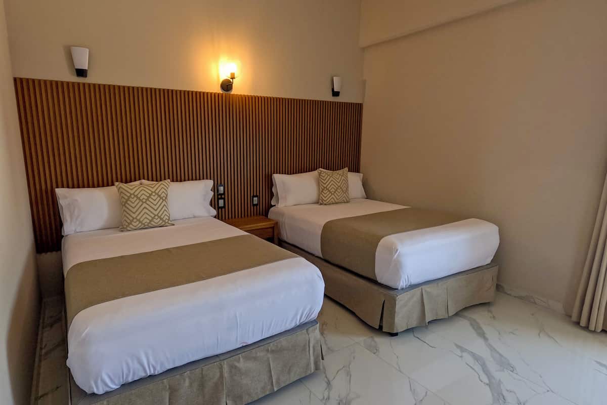 Una habitación de hotel con dos camas, cabecero de madera, suelo de mármol e iluminación en la pared.