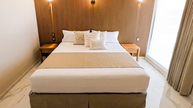 A hotel room with a large bed, wooden panel wall, two bedside tables, and a window with curtains. - Hotel JVD Inn - Tonalá Centro (Tonalá)