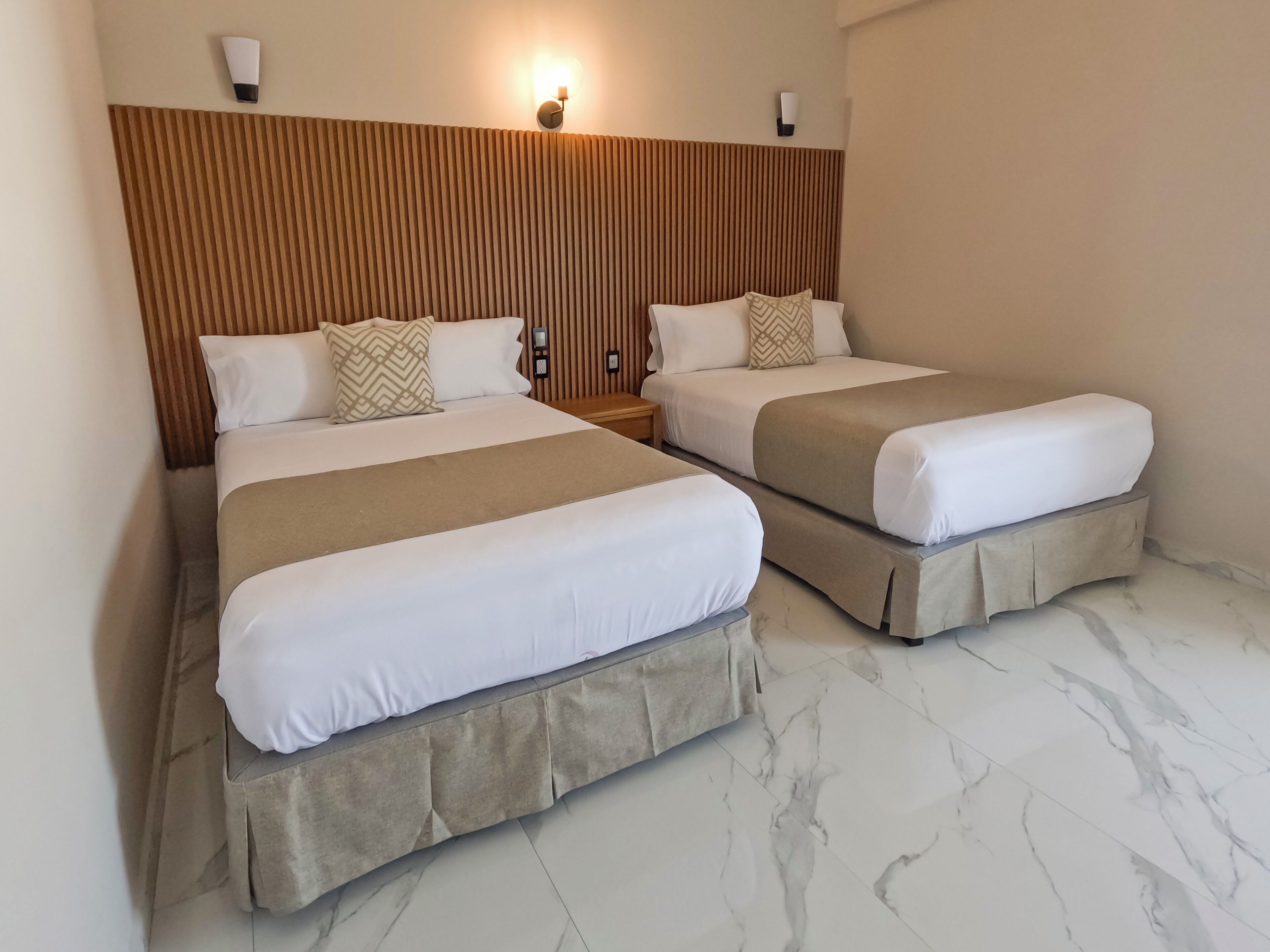 Dos camas individuales con cabeceras, una mesita de noche y una lámpara de pared en una habitación de hotel.