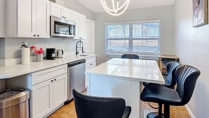 Une cuisine moderne avec des meubles blancs, un four à micro-ondes et un lave-vaisselle. On y trouve un coin repas avec une table et des chaises, ainsi qu’une fenêtre avec des stores.