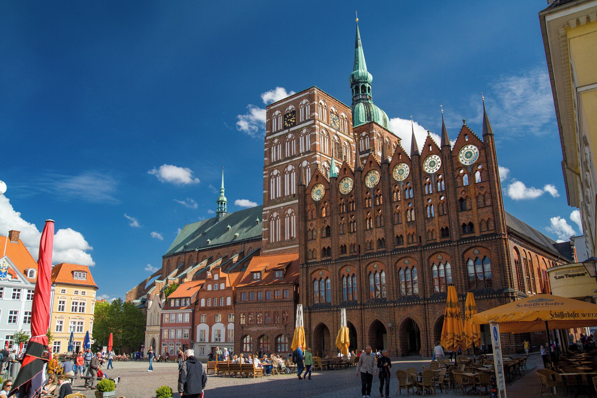En historisk byggnad med ett markant torn och en klocka, omgiven av ett torg med kullerstenar, utomhusservering och parasoller.