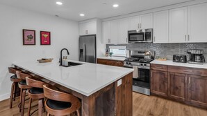 Una cocina moderna con armarios blancos, electrodomésticos de acero inoxidable, una isla central con taburetes y un salpicadero con diseño geométrico.