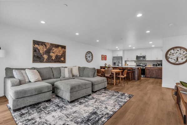 A modern living room with a sectional sofa, a dining area, and a kitchen with white cabinets and stainless steel appliances.